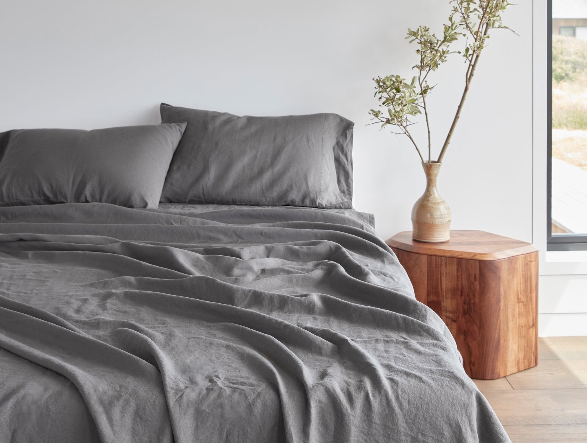 Coyuchi Organic Relaxed Linen Fitted Sheet in Slate – made bed beside bright window, showing colour in natural light. #colour_Slate