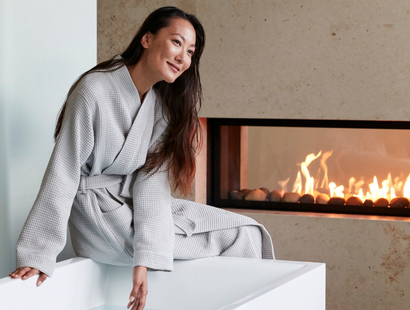 Coyuchi Organic Cotton Unisex Waffle Robe - model wearing in front of fireplace beside bath tub #colour_fog