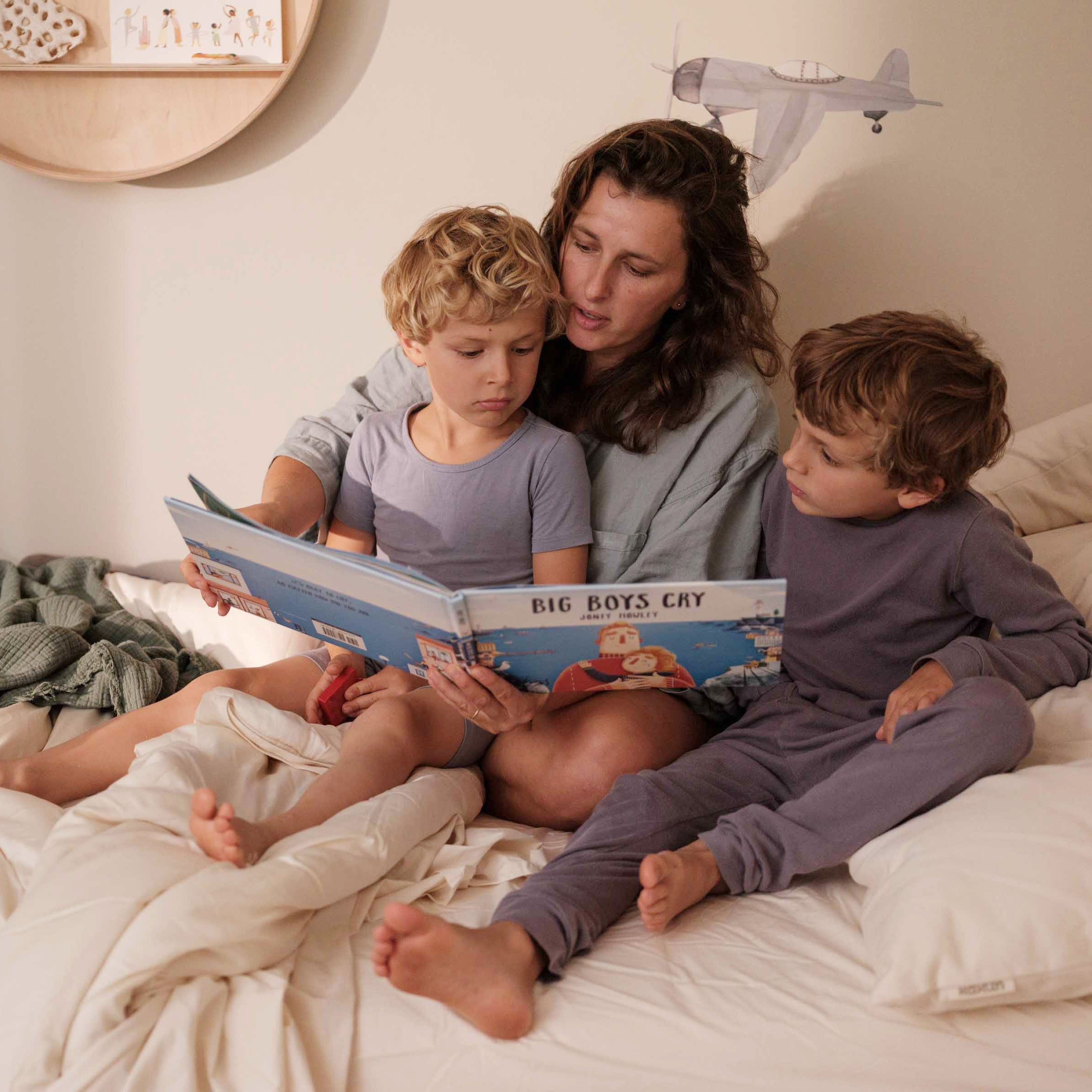 Woman reading a book to two children on a bed in a cozy room.